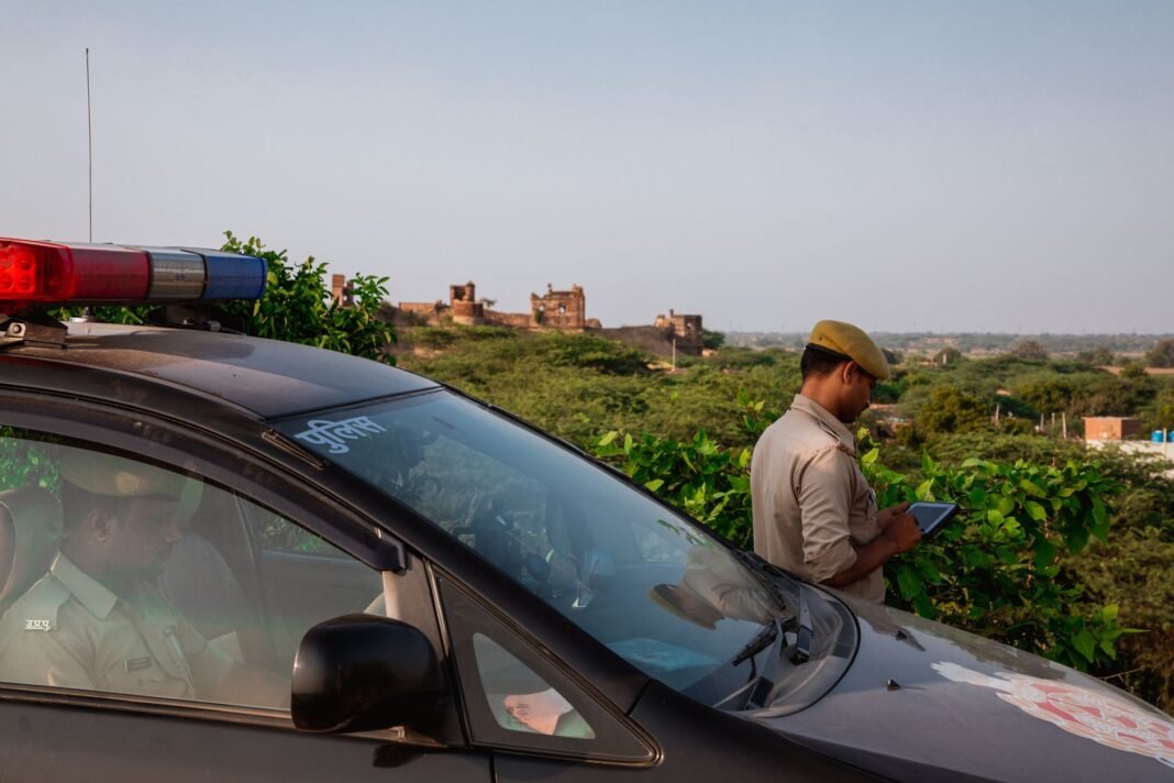 Three detained after video of minor girl being harassed in Jamui surfaces online Police officer using tablet beside patrol car in scenic outdoor setting.